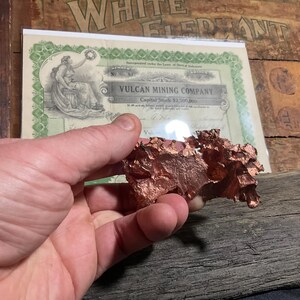 May include: A rough, reddish-brown copper specimen is held in a hand. Behind it is a vintage stock certificate from the Vulcan Mining Company. The certificate has green and white details and an illustration of a woman.