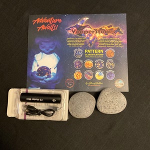 May include: Two gray rocks, a black charging cord, and a black charging brick in a clear plastic container. A card with the text "Adventure Awaits! Yooperstones" and a chart of different rock patterns is in the background.
