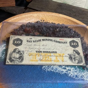 May include: A framed antique Bay State Mining Company ten-dollar bill, featuring portraits and ornate text. The bill is set against a blue and white wave design, with dark stones and resin in a wooden bowl.