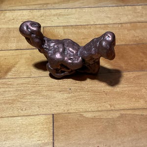 May include: A raw, metallic copper nugget with an irregular, organic shape. The surface features a deep, reddish-brown hue with a polished, reflective sheen and multiple rounded protrusions, creating a textured, uneven appearance throughout the piece.