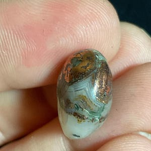 May include: An oval-shaped gemstone with a mix of colors, including white, brown, and green. The stone has a polished surface and appears to be held in a person's fingers. The gemstone has a unique pattern and texture.