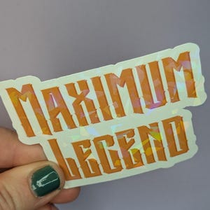 May include: A holographic sticker with the words "MAXIMUM LEGEND" in orange block letters. The sticker has a white border and a reflective, iridescent finish, creating a shimmering effect. The sticker is held against a light purple background.
