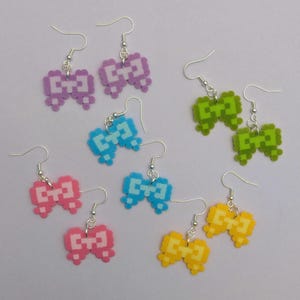 May include: Pixelated bow earrings in various colors: purple, green, blue, pink, and yellow. Each pair features a bow design made from small, square beads, with silver-colored ear hooks. A fun and colorful accessory.