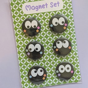 May include: A set of six black round magnets with white eyes and colorful star accents. The magnets are packaged on a green patterned card with the words "Magnet Set" in a purple font.