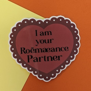 May include: A heart-shaped sticker with a scalloped edge and the text "I am your Roëmaance Partner" in black. The heart is burgundy with a white dotted border, set against a yellow and orange background.