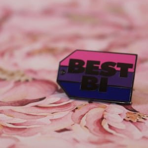 Puede incluir: Un pin de esmalte rosa, morado y azul con el texto "BEST BI" en letras negras.