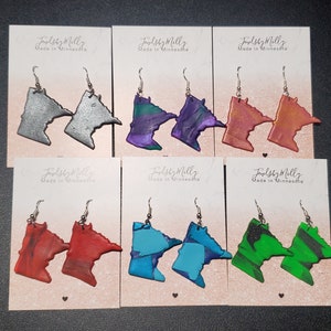 May include: Six pairs of Minnesota-shaped earrings on display cards. The earrings are made of polymer clay and come in a variety of colors, including silver, purple, orange, red, blue, and green.