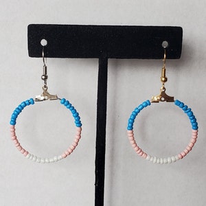 Puede incluir: Un par de pendientes de aro con un diseño de cuentas. Los pendientes presentan una combinación de colores de cuentas azules, rosas y blancas, dispuestas en un patrón circular. Los pendientes están suspendidos de ganchos plateados.