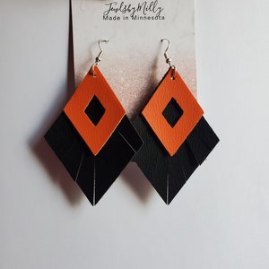 Puede incluir: Un par de pendientes de cuero negro y naranja con forma de diamante. Los pendientes tienen un detalle de flecos y un pequeño recorte cuadrado en el centro.