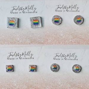 May include: Four pairs of stud earrings with a rainbow flag design. Two pairs are square, and two are round, all set in a silver glitter base. The text "Jewlsby Milly Made in Minnesota" is above each pair.