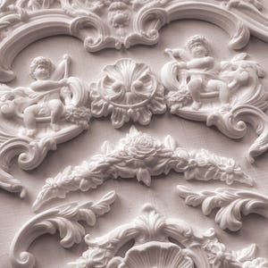 May include: White decorative plaster architectural elements. The image shows various ornate designs, including cherubs, floral garlands, and scrolled frames. The details are finely sculpted, creating a classical aesthetic. The pieces are likely for interior design or craft projects.