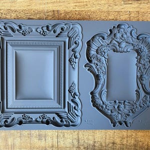 May include: A gray silicone mold featuring two ornate picture frame designs. The frames have detailed scrollwork and floral accents. The mold is labeled "FRAMES IOD".