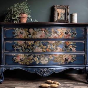May include: A navy blue three-drawer dresser with floral decals. The dresser has ornate handles and legs. The drawers are decorated with a floral pattern of pink, yellow, and orange flowers. The top of the dresser is a dark brown color.