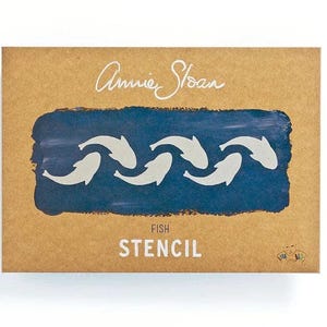 May include: A brown cardboard box featuring a blue painted rectangle with a white fish stencil design. The words "Annie Sloan" are written in cursive at the top, with "FISH STENCIL" printed below the design. The box is on a white background.