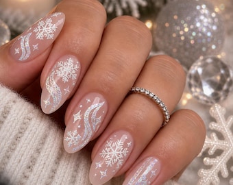 Puede incluir: Uñas en forma de almendra con una base nude transparente, decoradas con calcomanías de copos de nieve blancos y plateados iridiscentes, patrones de cintas onduladas y pequeños detalles de estrellas. Un anillo fino de metal plateado con piedras.