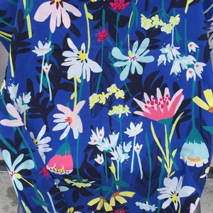 May include: A fabric featuring a vibrant royal blue background with a dense, colorful floral pattern. The design includes stylized flowers in shades of white, pale pink, bright yellow, light blue, and magenta, interspersed with dark navy green leaves.