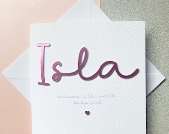 May include: A white card with the name "Isla" in pink script, with the words "welcome to the world baby girl" below. A small pink heart is at the bottom. The card is inside a white envelope, set against a pink and silver background.