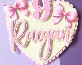 May include: A layered paper cake topper in the shape of a heart, with the name "Raegan" and the number "9" in pink. Two pink bows are attached to the heart. The topper is on a white stick.
