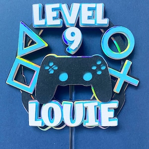 Op de afbeelding: Een caketopper met een gamingthema. Het bevat de woorden "LEVEL 9 LOUIE" in witte, gelaagde tekst. Een zwarte gamecontroller staat in het midden, omringd door blauwe en holografische gamesymbolen. De topper staat op een doorzichtige stok.
