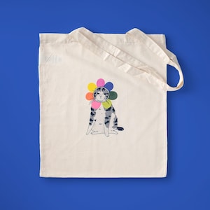 Puede incluir: Bolso tote de lona color crema con una ilustración de un gato gris y blanco con una corona de flores de globos de colores. El bolso tiene dos asas y está sobre un fondo azul.