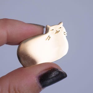 May include: A gold-coloured cat-shaped pin. The pin is smooth and rounded, with etched details for the eyes, nose, and whiskers. The cat is in a relaxed pose, with its eyes closed. The pin is held between two fingers.