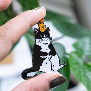 May include: A black and white cat enamel pin with a stack of orange fruit on its head. The cat is sitting with its legs out and arms crossed. The pin is held by a person's fingers in front of a green leafy background.