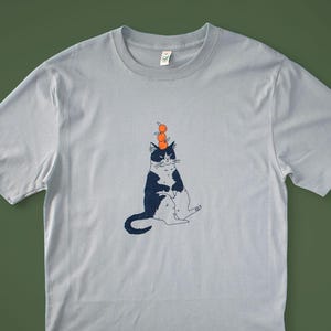 May include: Light blue t-shirt featuring a whimsical illustration of a black and white cat balancing three orange spheres on its head. The cat is sitting with its legs extended. The shirt has a crew neck and short sleeves.