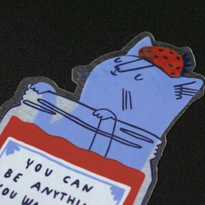 May include: A blue cat with a strawberry on its head is inside a glass jar filled with red jam. The jar has a white label with the text "You can be anything you wa".