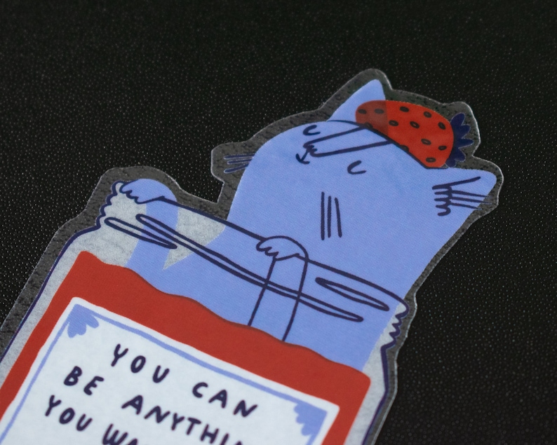 May include: A blue cat with a strawberry on its head is inside a glass jar filled with red jam. The jar has a white label with the text "You can be anything you wa".