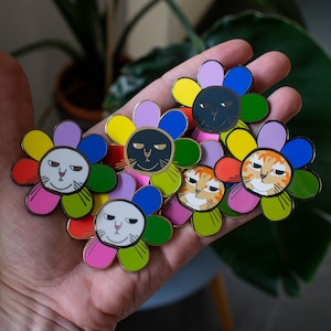 Può includere: Un set di sei spille smaltate con disegni di fiori colorati con volti di gatto. Ogni spilla ha uno schema di colori e un'espressione di gatto diversi.