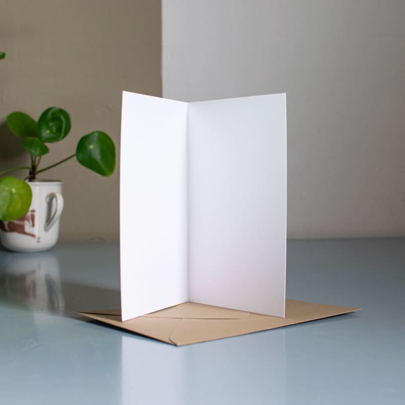 May include: A blank white card folded in half and placed on a brown envelope. The card is sitting on a light blue surface.