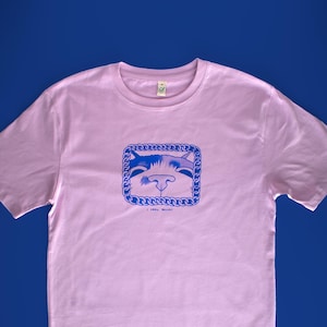 以下が含まれることがあります： 水色の猫の顔のグラフィックがチェーンのような枠の中に入った、薄いピンク色のTシャツ。「I AM MEOW」の文字がグラフィックの下にあります。Tシャツは半袖で、クルーネックです。