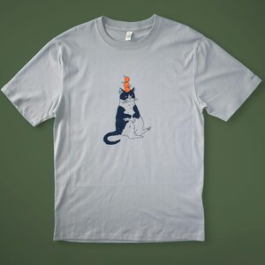 May include: Light gray t-shirt featuring a whimsical illustration of a black and white cat balancing three orange spheres on its head. The cat is seated with its legs crossed. The shirt is made of a soft material.