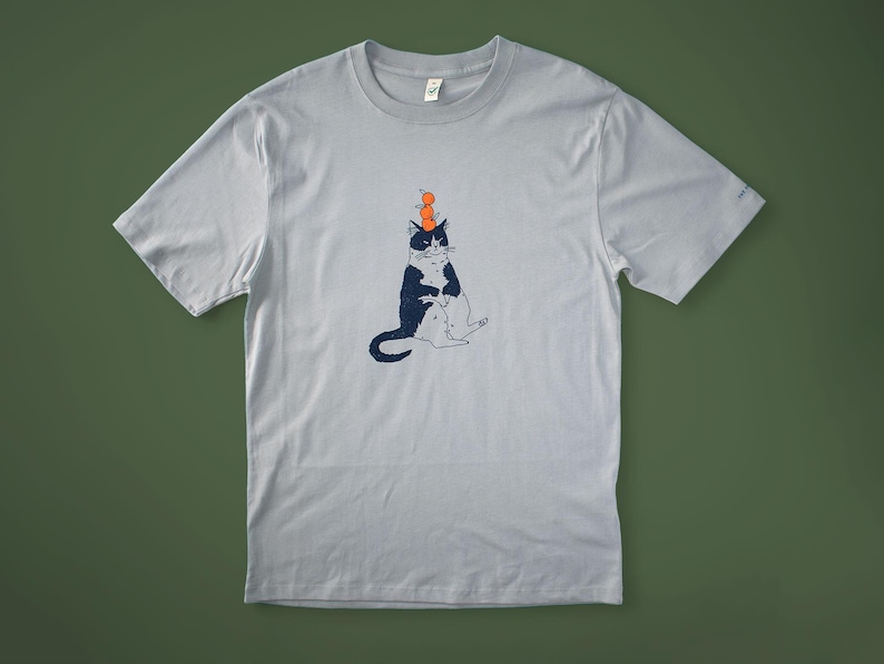 May include: Light gray t-shirt featuring a whimsical illustration of a black and white cat balancing three orange spheres on its head. The cat is seated with its legs crossed. The shirt is made of a soft material.