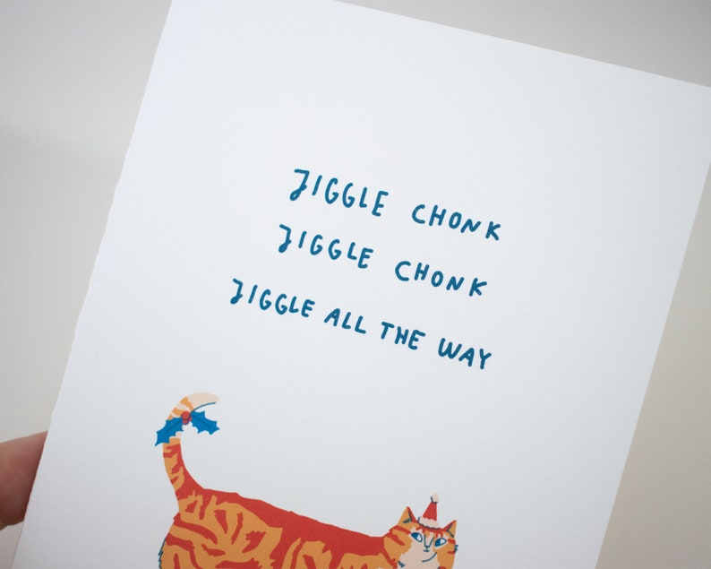May include: A white card with the text "Jiggle Chonk Jiggle Chonk Jiggle All The Way" in blue handwriting. A cartoon orange and white cat with a red Santa hat and holly on its tail is at the bottom of the card.
