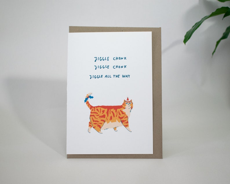 May include: A greeting card with a cartoon orange and red tabby cat wearing a blue and white party hat. The text on the card reads "Jiggle Chonk Jiggle Chonk Jiggle All The Way."