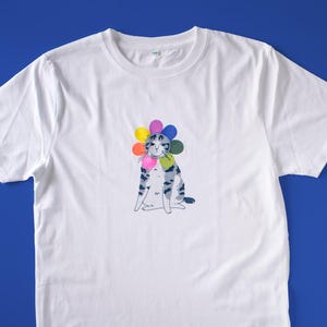 May include: White t-shirt featuring a whimsical illustration of a cat wearing a colorful flower crown. The cat has a gray and blue striped pattern. The shirt has short sleeves and a crew neck. The text "THE FRUIT" is printed on the sleeve.