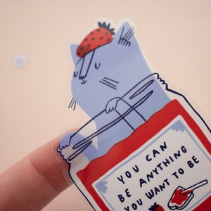 May include: A blue cat wearing a red strawberry hat is inside a clear jar filled with red jam. The jar has a white label with the text "You can be anything you want to be".