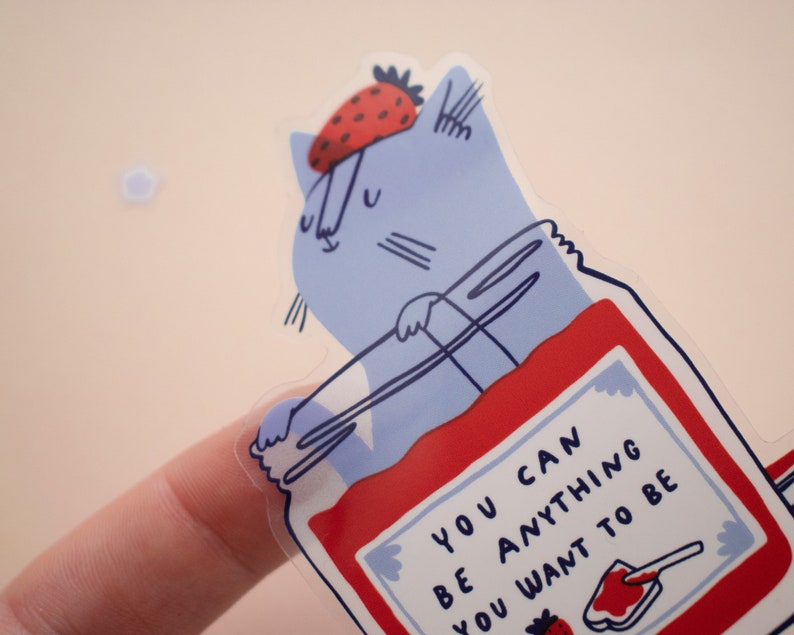 May include: A blue cat wearing a red strawberry hat is inside a clear jar filled with red jam. The jar has a white label with the text "You can be anything you want to be".