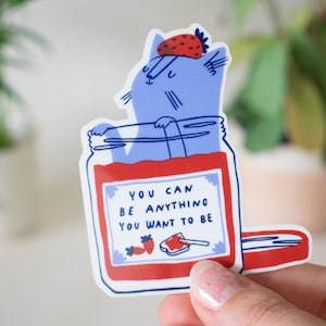 May include: A blue cat wearing a strawberry hat is inside a red jar with the text "You can be anything you want to be".