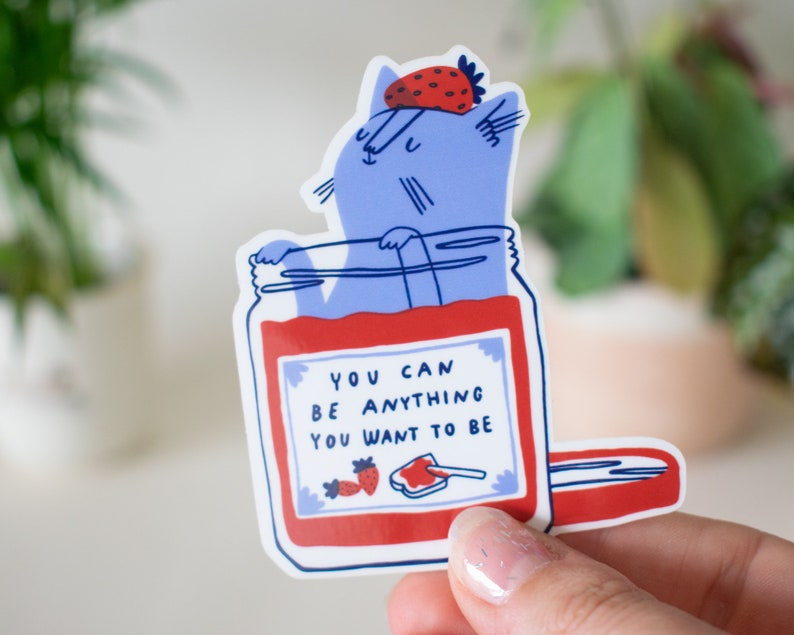 May include: A blue cat wearing a strawberry hat is inside a red jar with the text "You can be anything you want to be".