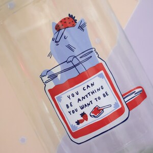 May include: A glass jar with a blue cat wearing a strawberry on its head. The jar is filled with red jam and has a white label with the text "You can be anything you want to be".