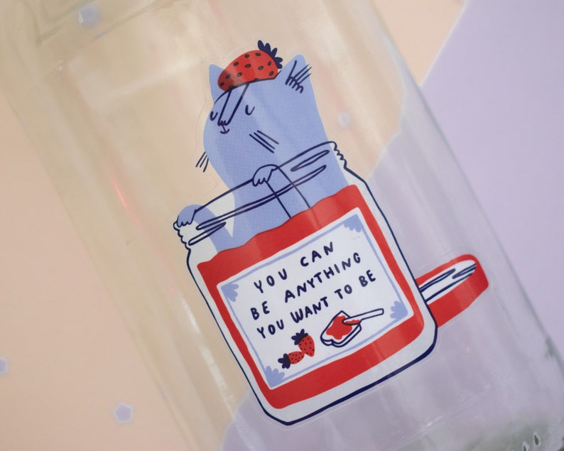 May include: A glass jar with a blue cat wearing a strawberry on its head. The jar is filled with red jam and has a white label with the text "You can be anything you want to be".