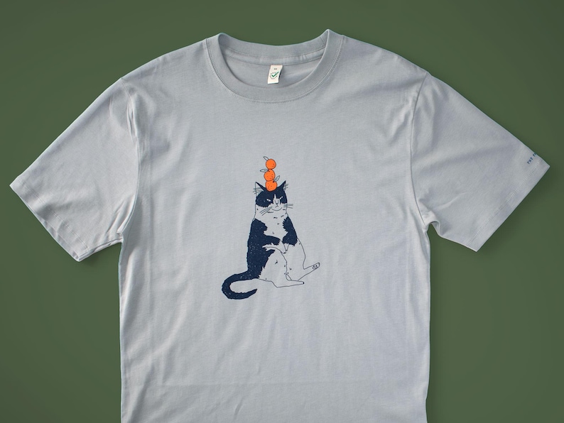 May include: Light blue t-shirt featuring a whimsical illustration of a black and white cat balancing three orange spheres on its head. The cat is sitting with its legs extended. The shirt has a crew neck and short sleeves.