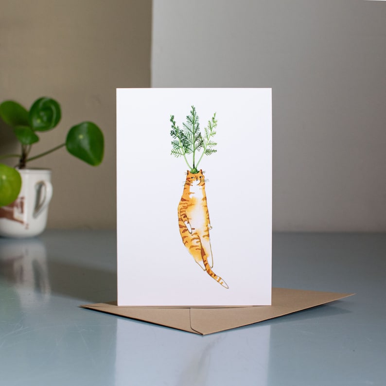 May include: A greeting card featuring a watercolor illustration of an orange tabby cat with green leafy stems growing out of its head, resembling a carrot.