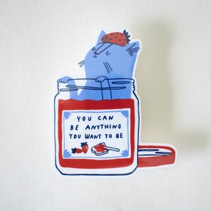 May include: A blue cat wearing a strawberry on its head is inside a glass jar filled with red jam. The jar has a white label with the text "You can be anything you want to be".