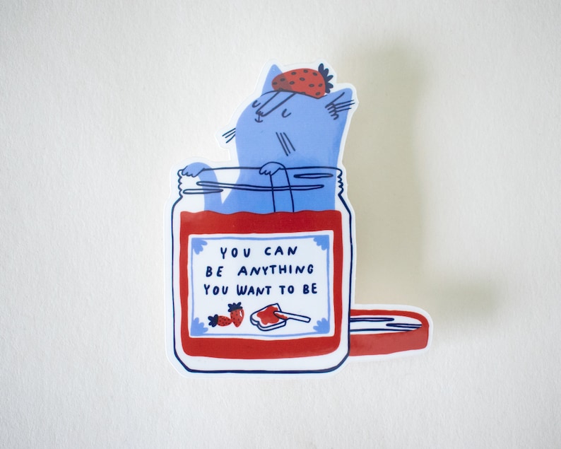 May include: A blue cat wearing a strawberry on its head is inside a glass jar filled with red jam. The jar has a white label with the text "You can be anything you want to be".