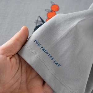 May include: A light gray t-shirt with a graphic design of a cat and oranges. The text "THE PRINTED CAT" is printed in blue on the sleeve. The shirt is made of a soft, comfortable material.