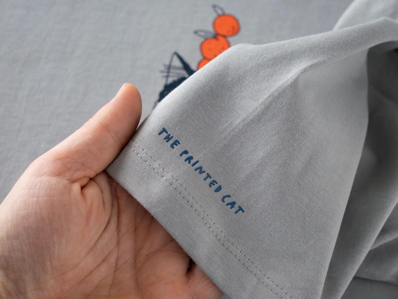 May include: A light gray t-shirt with a graphic design of a cat and oranges. The text "THE PRINTED CAT" is printed in blue on the sleeve. The shirt is made of a soft, comfortable material.