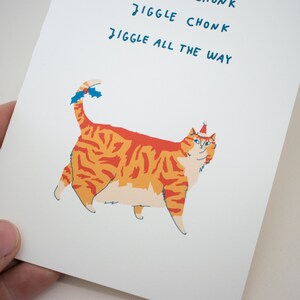 May include: A greeting card featuring a cartoon orange tabby cat wearing a red Santa hat with a blue trim. The cat has a white chest and belly. The text on the card reads "Jiggle Chonk Jiggle All The Way".